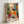 May include: Colorful Matisse-style art poster in light wooden frame leaning against white wall. Shows vibrant interior scene with woman in yellow-orange dress sitting at table with fruit bowl, teacup, and red flowers in blue vase. Features bold fauve colors - greens, oranges, reds, blues - with decorative patterns and window view. Artist name "MATISSE" printed at bottom in black letters.