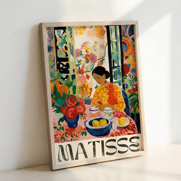 May include: Colorful Matisse-style art poster in light wooden frame leaning against white wall. Shows vibrant interior scene with woman in yellow-orange dress sitting at table with fruit bowl, teacup, and red flowers in blue vase. Features bold fauve colors - greens, oranges, reds, blues - with decorative patterns and window view. Artist name "MATISSE" printed at bottom in black letters.