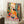 May include: Framed colorful poster featuring a woman in yellow dress sitting at a table with fruit bowl and teacups, surrounded by vibrant tropical plants and flowers in Matisse's distinctive style. The artwork shows bold oranges, greens, and blues with "MATISSE" text at bottom. Frame leans against white wall next to coffee cup on wooden block and cream textured blanket on fluffy white rug.