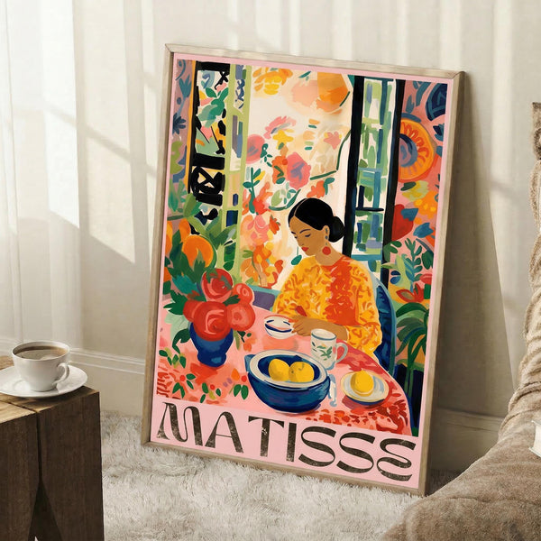 May include: Framed colorful poster featuring a woman in yellow dress sitting at a table with fruit bowl and teacups, surrounded by vibrant tropical plants and flowers in Matisse's distinctive style. The artwork shows bold oranges, greens, and blues with "MATISSE" text at bottom. Frame leans against white wall next to coffee cup on wooden block and cream textured blanket on fluffy white rug.