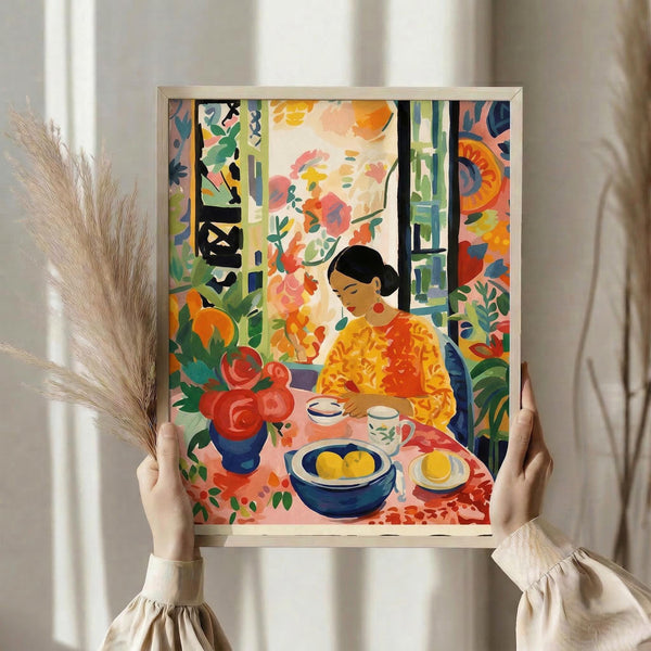 May include: Hands holding a framed colorful artwork depicting a woman in yellow clothing sitting at a table with fruit and flowers. The painting features vibrant oranges, greens, and blues in an expressive style with floral patterns and interior elements. Natural lighting creates soft shadows on neutral curtains in the background, with dried wheat stems visible on the left side.