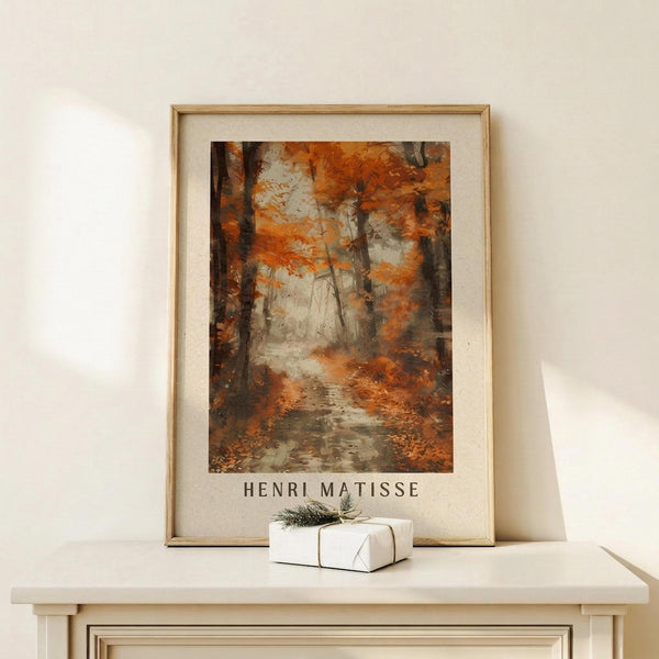 May include: Framed poster print featuring an autumn forest scene with orange and brown foliage, dark tree trunks, and a misty pathway. The artwork shows painterly brushstrokes in warm earth tones with "HENRI MATISSE" text below. Natural wood frame with white matting, displayed on light wooden surface beside small white wrapped gift with twine and evergreen sprig against cream wall.