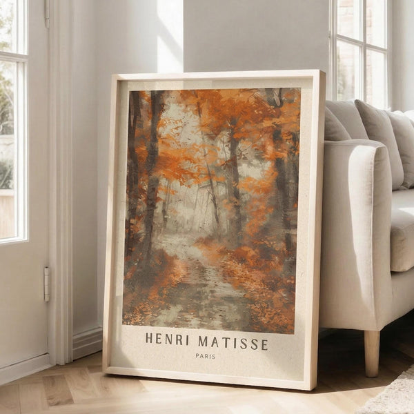 May include: Framed art print showing an autumn forest scene with orange and brown foliage, bare trees, and a misty path through woods. The poster features "HENRI MATISSE PARIS" text at bottom and sits in a light wood frame, leaning against a white wall next to a beige upholstered sofa in a bright, minimalist living room with large windows.