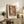 May include: Framed autumn forest artwork with orange and brown foliage displayed on rustic wooden sideboard alongside fern plant, ceramic vase, table lamp, and leather chair in warm-toned living room setting