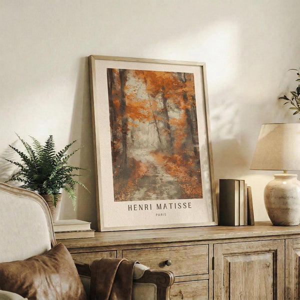 May include: Framed autumn forest artwork with orange and brown foliage displayed on rustic wooden sideboard alongside fern plant, ceramic vase, table lamp, and leather chair in warm-toned living room setting