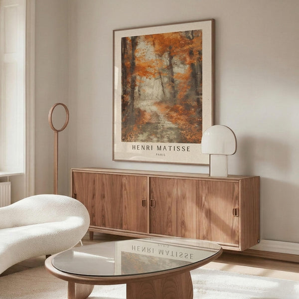 May include: Henri Matisse forest poster print with autumn orange and brown tones displayed above a wood credenza in minimalist living room. White curved chair, glass coffee table, and modern lamp complete the neutral decor scene with warm natural lighting.