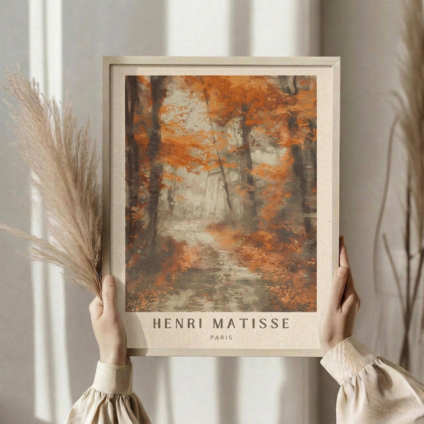 May include: Hands holding a framed art print featuring an autumn forest scene with orange and brown foliage, trees, and a winding path. The print has "Henri Matisse Paris" text at the bottom and is displayed in a light wood frame. Dried pampas grass stems are visible to the left, with soft natural lighting and neutral curtains in the background creating a warm, minimalist home setting.