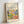 May include: Framed art poster with cream background displaying colorful abstract forest scene in Matisse's style, featuring vibrant green foliage, purple and black tree trunks, yellow and orange circular shapes, and blue botanical elements. Text reads "Discover the Beauty of Matisse's Art" above the artwork. Natural wood frame leans against white wall on light surface, casting soft shadows.