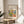 May include: Framed art poster with colorful abstract garden scene featuring vibrant yellows, greens, blues and purples in Matisse's distinctive cut-out style. Text reads "Discover the Beauty of Matisse's Art" above the artwork. Thin wooden frame displayed on round light wood side table next to beige upholstered chair. Natural sunlight creates geometric shadows on white wall. Green plant leaves visible in corner, creating modern minimalist home decor setting.