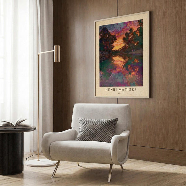 May include: Henri Matisse poster print in light wood frame hanging on wood-paneled wall above beige upholstered armchair. The artwork shows a colorful landscape with trees reflected in water, featuring vibrant purples, oranges, and greens in an impressionistic style. Modern reading corner with geometric patterned pillow, brass floor lamp, and round side table with open book.