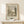 May include: Framed vintage botanical illustration print leaning against white wall on light wooden surface. Black and white etching shows tranquil park scene with trees, water, and small structure. Aged cream matting with text details below artwork. Small white wrapped gift box with natural twine and greenery sprig positioned in front. Soft natural lighting creates gentle shadows on minimalist display.