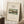 May include: Framed art print leaning against white wall on textured cream rug, featuring pastoral landscape with buildings and trees in muted greens and earth tones. Light wood frame, cream matting with black text above and below artwork. Coffee cup on wooden block nearby, cozy room setting with natural window light and beige throw blanket visible.