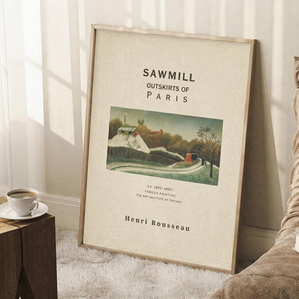 May include: Framed art print leaning against white wall on textured cream rug, featuring pastoral landscape with buildings and trees in muted greens and earth tones. Light wood frame, cream matting with black text above and below artwork. Coffee cup on wooden block nearby, cozy room setting with natural window light and beige throw blanket visible.