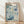 May include: Framed poster with blue and white floral design featuring abstract blooms, circles, and organic patterns in various shades of blue against cream background. Text reads "YAYOI KUSAMA" at top with Japanese characters and "1972" and "MUSEUM OF MODERN ART NEW YORK" at bottom. Light wood frame sits against white wall on wooden floor with soft natural lighting and shadows.