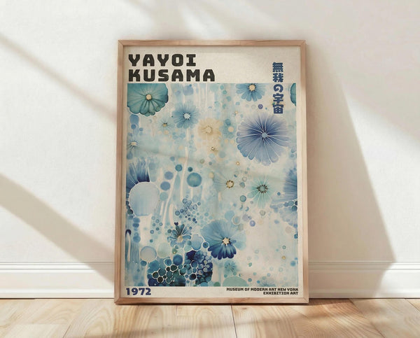 May include: Framed poster with blue and white floral design featuring abstract blooms, circles, and organic patterns in various shades of blue against cream background. Text reads "YAYOI KUSAMA" at top with Japanese characters and "1972" and "MUSEUM OF MODERN ART NEW YORK" at bottom. Light wood frame sits against white wall on wooden floor with soft natural lighting and shadows.