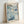 May include: Framed poster print leaning against white wall featuring abstract floral design in blue and teal tones with scattered circular patterns. Text reads "YAYOI KUSAMA" at top with Japanese characters and "1972" at bottom. Light wood frame surrounds cream-colored paper with watercolor-style artwork resembling flowers and dots in various shades of blue, green, and white.