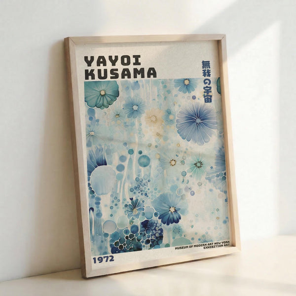 May include: Framed poster print leaning against white wall featuring abstract floral design in blue and teal tones with scattered circular patterns. Text reads "YAYOI KUSAMA" at top with Japanese characters and "1972" at bottom. Light wood frame surrounds cream-colored paper with watercolor-style artwork resembling flowers and dots in various shades of blue, green, and white.