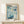May include: Framed art print in light wood frame displaying abstract floral design in blue and teal tones with scattered circles and geometric flower shapes. Features "YAYOI KUSAMA" text at top with Japanese characters and "1972" at bottom. Poster sits on light wooden surface next to small wrapped white gift box with twine and dried sprig against cream wall.