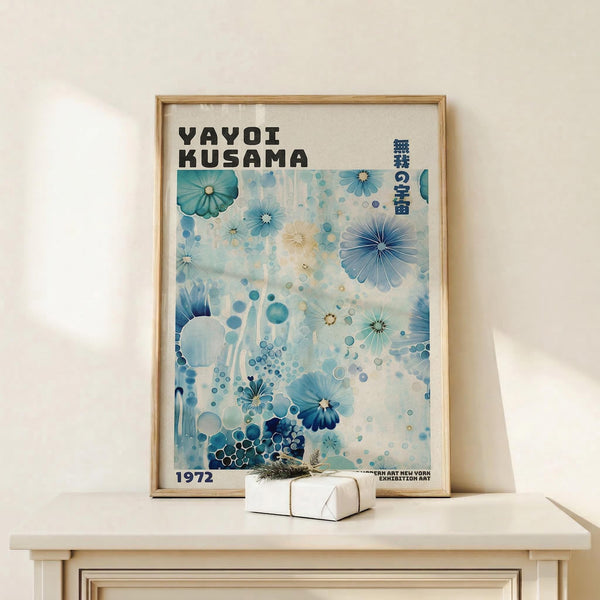 May include: Framed art print in light wood frame displaying abstract floral design in blue and teal tones with scattered circles and geometric flower shapes. Features "YAYOI KUSAMA" text at top with Japanese characters and "1972" at bottom. Poster sits on light wooden surface next to small wrapped white gift box with twine and dried sprig against cream wall.