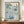 May include: Framed exhibition poster featuring blue watercolor floral patterns and polka dots with "YAYOI KUSAMA" text and "1972" date, displayed on wooden credenza next to white ceramic vase and stacked books in room with cream walls and white trim molding.