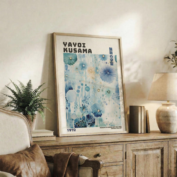 May include: Framed Yayoi Kusama museum poster with blue floral and polka dot design from 1972, displayed on rustic wooden sideboard next to fern plant, ceramic vases, and books. Light wooden frame against white wall in modern living room with leather chair visible in foreground.