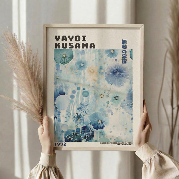May include: Hands holding a framed art print featuring blue watercolor flowers and circles in various shades, with "YAYOI KUSAMA" text at top and "1972" at bottom. Light wooden frame against neutral curtained background with dried pampas grass visible on left side. Soft natural lighting creates gentle shadows.