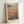 May include: Framed poster print featuring Yayoi Kusama's polka dot pumpkin installation artwork in warm orange and yellow tones against green background, displayed in light wooden frame leaning against white wall corner. Japanese text appears at top and bottom of vintage-style museum exhibition poster.