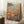 May include: Framed poster featuring Yayoi Kusama's polka dot artwork with yellow, orange, and pink circular forms on green background, displayed in light wooden frame leaning against white wall next to beige textured blanket and wooden side table with white coffee cup and saucer on white fluffy rug