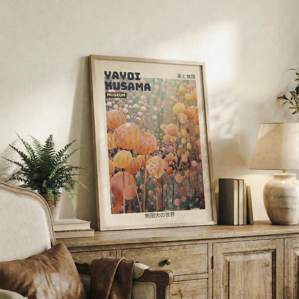 May include: Framed art print featuring colorful polka-dotted spherical sculptures in orange, pink, and yellow tones with Japanese text, displayed on rustic wooden console table alongside fern plant, ceramic vase, and stacked books in warm neutral living room setting with beige armchair.