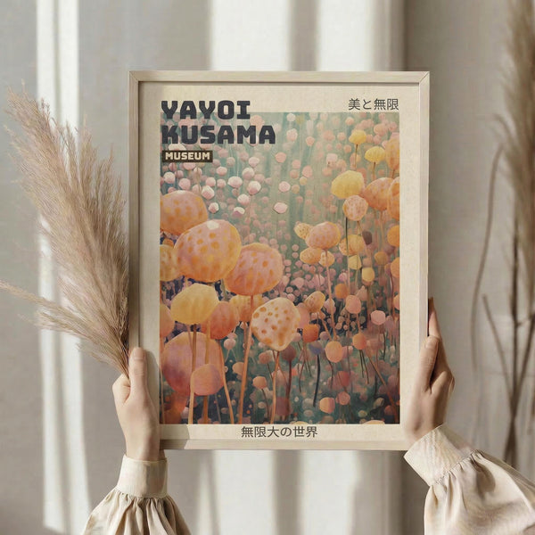 May include: Hands holding a framed art print featuring abstract polka-dotted spherical forms in warm orange, pink and yellow tones against a muted background. The poster displays "YAYOI KUSAMA MUSEUM" text with Japanese characters. Person wears a cream-colored ruffled sleeve top while holding dried pampas grass stems. Light wooden frame against soft neutral curtained background creates an aesthetic home decor styling scene.