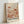May include: Framed art print leaning against white wall showing abstract botanical illustration in warm coral, peach and cream tones. Features stylized mushrooms, flowers, and organic shapes with polka dot patterns and scattered circular elements. Text reads "YAYOI KUSAMA" and "KYOTO" in black letters at bottom with Japanese characters. Light wooden frame casts soft shadow on textured white surface.
