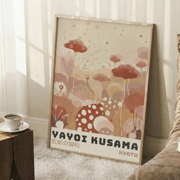 May include: Framed art print leaning against white wall in cozy room setting with coffee cup on wooden block and knit blanket. The poster features abstract botanical illustration in warm coral, peach and cream tones with polka dot mushrooms, fan-shaped plants, and organic forms. Text reads "YAYOI KUSAMA" and Japanese characters with "KYOTO" below in modern typography on cream background.
