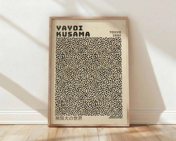 May include: Framed exhibition poster with cream background featuring Yayoi Kusama's name in bold black letters, dense pattern of small black polka dots covering most of the surface, Japanese text at bottom, "TOKYO 1992" in upper right corner, displayed in light wood frame leaning against white textured wall on wooden floor with natural lighting creating soft shadows.