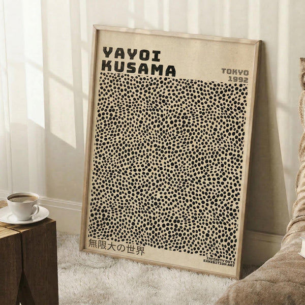 May include: Framed art print leaning against white wall featuring black polka dot pattern on cream background with "YAYOI KUSAMA" text and "TOKYO 1992" in upper corner, Japanese text at bottom. Light wood frame, displayed in cozy room setting with coffee cup on wooden block side table and soft textured throw blanket visible.