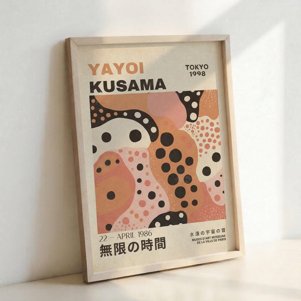 Framed exhibition poster featuring abstract organic shapes in coral, peach, black and cream with polka dot patterns. The artwork displays "YAYOI KUSAMA" prominently at top with "TOKYO 1998" in upper right corner. Japanese text appears at bottom along with "22 - APRIL 1986" and museum information. The poster sits in a light wooden frame against a white wall, casting soft shadows. The design showcases Kusama's signature spotted motifs in flowing, curved forms with varying dot