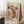 May include: Framed Yayoi Kusama exhibition poster leaning against white wall corner on white fluffy rug. Features abstract polka dot design in coral pink, orange, black and white organic shapes. Text shows "YAYOI KUSAMA" "TOKYO 1998" and Japanese characters. Light wood frame, styled with small white coffee cup on wooden block and beige knit blanket. Soft natural lighting creates gentle shadows on textured wall.