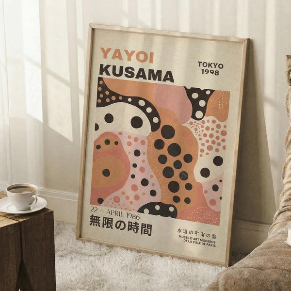 May include: Framed Yayoi Kusama exhibition poster leaning against white wall corner on white fluffy rug. Features abstract polka dot design in coral pink, orange, black and white organic shapes. Text shows "YAYOI KUSAMA" "TOKYO 1998" and Japanese characters. Light wood frame, styled with small white coffee cup on wooden block and beige knit blanket. Soft natural lighting creates gentle shadows on textured wall.