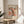 May include: Framed art poster featuring abstract organic shapes in coral, peach, and black with polka dot patterns, displayed on a light wood round side table next to a beige upholstered chair. Natural sunlight streams across the white wall behind, with green plant leaves visible in the corner. The poster shows exhibition text in both English and Japanese characters.