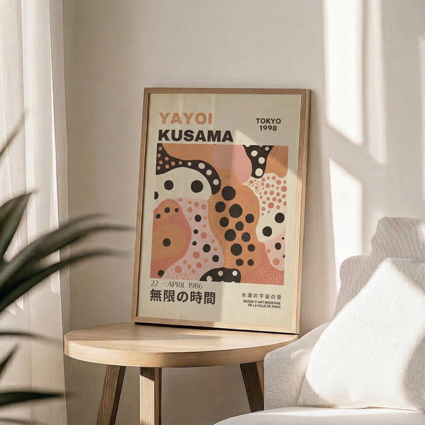 May include: Framed art poster featuring abstract organic shapes in coral, peach, and black with polka dot patterns, displayed on a light wood round side table next to a beige upholstered chair. Natural sunlight streams across the white wall behind, with green plant leaves visible in the corner. The poster shows exhibition text in both English and Japanese characters.