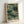 May include: Framed art print leaning against white wall showing abstract botanical illustration in muted green, sage, and cream tones with delicate flowering branches, dotted textures, and organic shapes. Light wooden frame surrounds artwork featuring "YAYOI KUSAMA TOKYO" text at bottom with Japanese characters below.