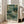 May include: Framed botanical art print leaning against white wall beside beige couch in bright living room. Artwork features layered green foliage patterns with cream flowers and dotted textures in nature-inspired design. Light wood frame, approximately 2-3 feet tall, positioned on light hardwood floors near large windows with natural sunlight streaming in.