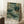 May include: Framed poster with abstract botanical artwork featuring flowing green and teal organic shapes, delicate white flowers, and dotted patterns in Kusama's signature style. Text reads "YAYOI KUSAMA" with Japanese characters and "TOKYO" below. Light wood frame leans against white wall next to wooden stool with coffee cup, cream textured rug visible.