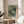 May include: Framed art print featuring green botanical illustration with delicate wildflowers, dotted patterns, and organic shapes in sage and cream tones. Print displays "YAYOI KUSAMA" and "TOKYO" text below artwork. Wooden frame sits on round light wood side table next to beige upholstered chair. Soft natural lighting creates shadows on neutral wall background with plant leaves visible in corner.