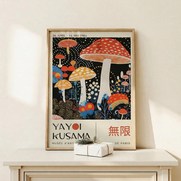 May include: A framed poster featuring colorful mushrooms on a black starry background with white polka dots. The artwork shows red, orange, and yellow spotted mushrooms with detailed gills, surrounded by small white dots resembling stars or spores. Text reads "YAYOI KUSAMA" and "MUSÉE d'ART DE PARIS" with Japanese characters. The wooden frame sits on a light wooden shelf next to a small white wrapped gift with natural twine and greenery.