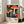 May include: Large framed poster leaning against white wall in bright living room with gray sofa. Artwork features vibrant mushrooms in red, orange, and yellow with white polka dots against black starry background, with blue botanical elements. Text shows "YAYOI KUSAMA" and museum information below the whimsical illustration. Natural lighting creates soft shadows on wooden floor.