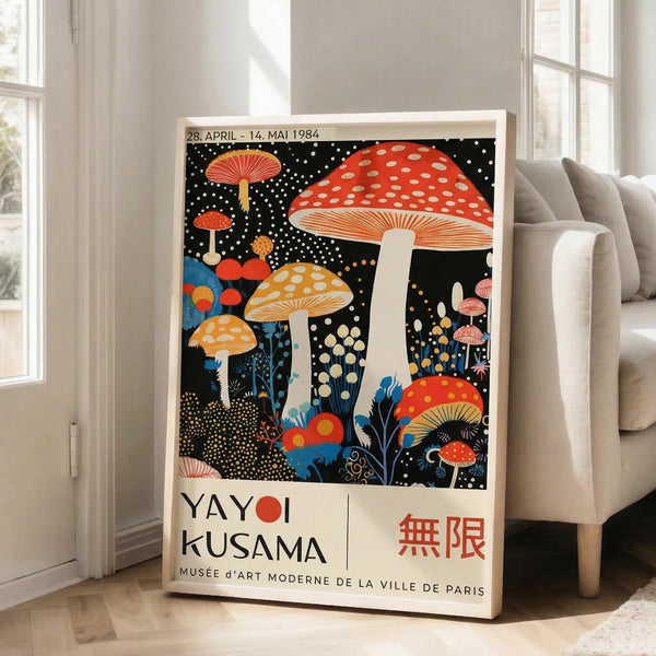 May include: Large framed poster leaning against white wall in bright living room with gray sofa. Artwork features vibrant mushrooms in red, orange, and yellow with white polka dots against black starry background, with blue botanical elements. Text shows "YAYOI KUSAMA" and museum information below the whimsical illustration. Natural lighting creates soft shadows on wooden floor.