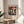 May include: Framed museum exhibition poster featuring colorful mushrooms in red, orange, and yellow against black starry background, displayed on round wooden side table next to beige upholstered chair. Natural lighting creates soft shadows across neutral wall. Poster shows Japanese text and museum details at bottom in clean typography.