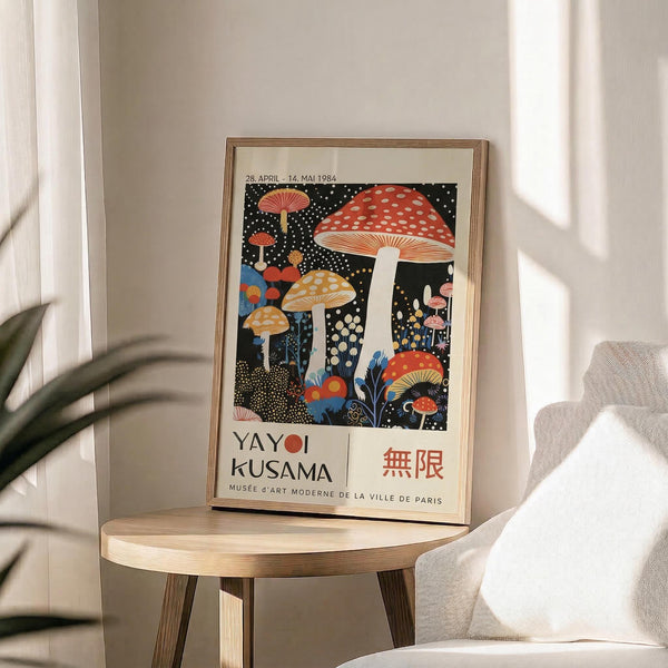 May include: Framed museum exhibition poster featuring colorful mushrooms in red, orange, and yellow against black starry background, displayed on round wooden side table next to beige upholstered chair. Natural lighting creates soft shadows across neutral wall. Poster shows Japanese text and museum details at bottom in clean typography.