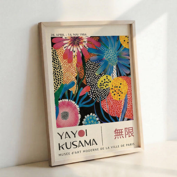 Framed exhibition poster featuring vibrant floral artwork with large stylized flowers in pink, blue, and yellow against dotted backgrounds in pop art style. The poster displays "YAYOI KUSAMA" and "MUSÉE D'ART MODERNE DE LA VILLE DE PARIS" text at bottom with exhibition dates. Light wood frame leans against white wall, showing the colorful botanical design with halftone dot patterns throughout.