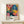 May include: Framed exhibition poster featuring vibrant floral artwork with colorful flowers in pink, blue, yellow, and orange against black background with polka dot patterns. Text shows "YAYOI KUSAMA" and "MUSEE d'ART DE PARIS" with dates "28. APRIL - 14. MAI 1984". Light wooden frame sits on white wooden surface next to small wrapped gift box with dried botanicals on top, against cream colored wall.
